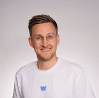 Freundlicher Mann mit Brille und weißem T-Shirt, lächelnd, vor neutralem Hintergrund.